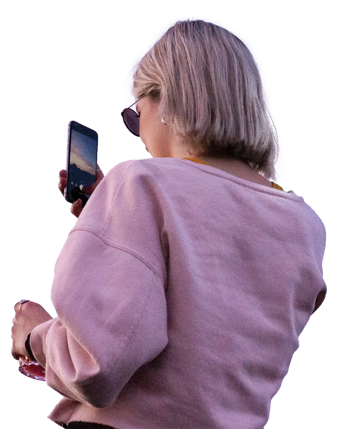 A woman stands outside with her back facing the viewer holding her phone depicting a sunset. She is wearing glasses, earrings and a pink sweater.