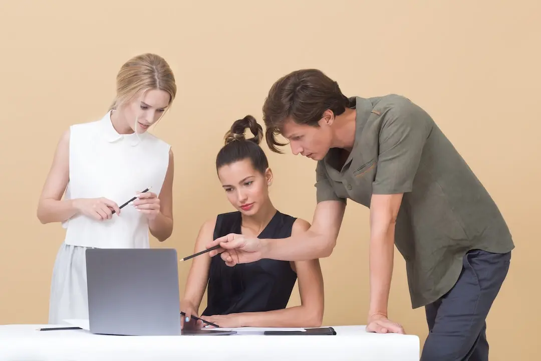 Three people stand around an unbranded laptop in front of a blank yellow background brandishing black pencils. The masculine indivdual on the right pokes at the laptop with his pencil while the others display a more passive demeanor, pencils at rest. The mood is introspective.