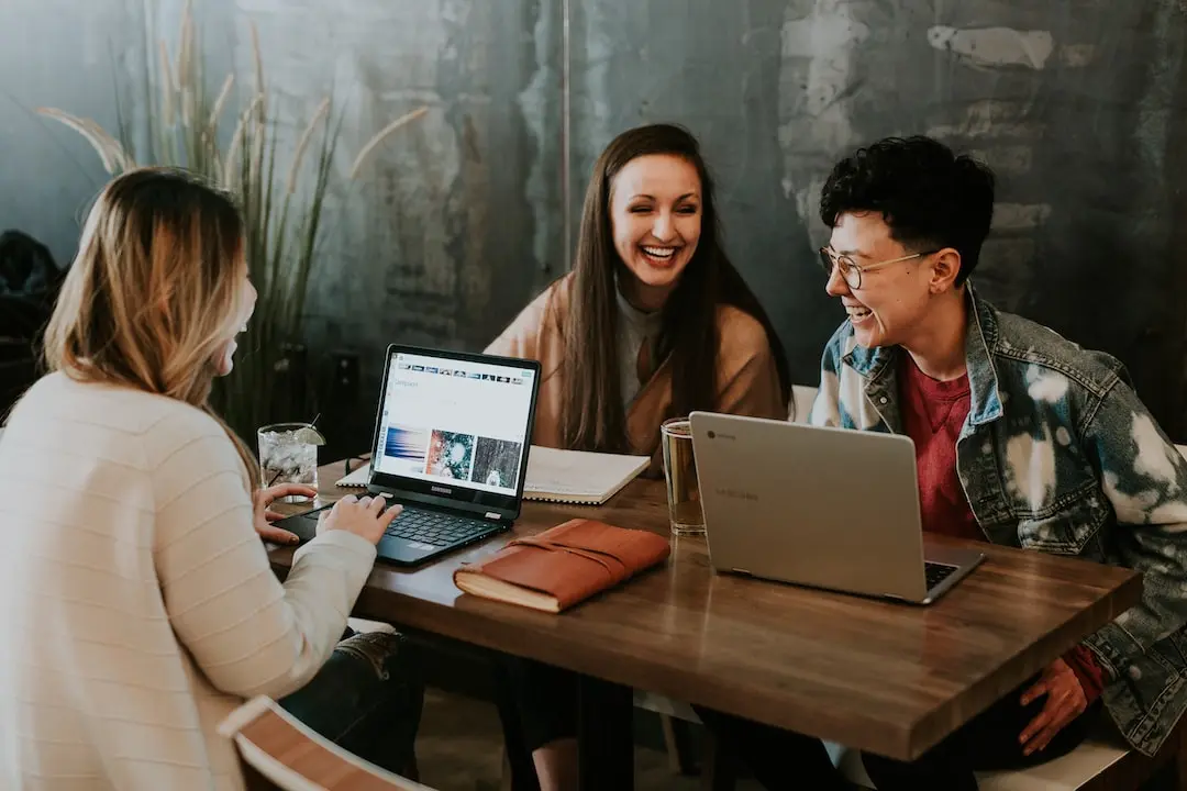 Three people sit around a table laughing at a shared happy thought. Items on the table are alcoholic drinks, laptops, notebooks and journals.